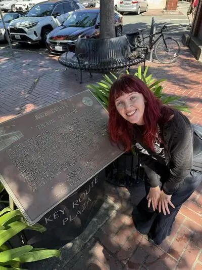 Kyra Eyerman posing beside the Key Route Train Station plaque in Oakland's Key Route Plaza. The plaque commemorates the arrival of the first Key Route electric train in 1904 and the station's role in East Bay transportation until the line's final run in 1958.