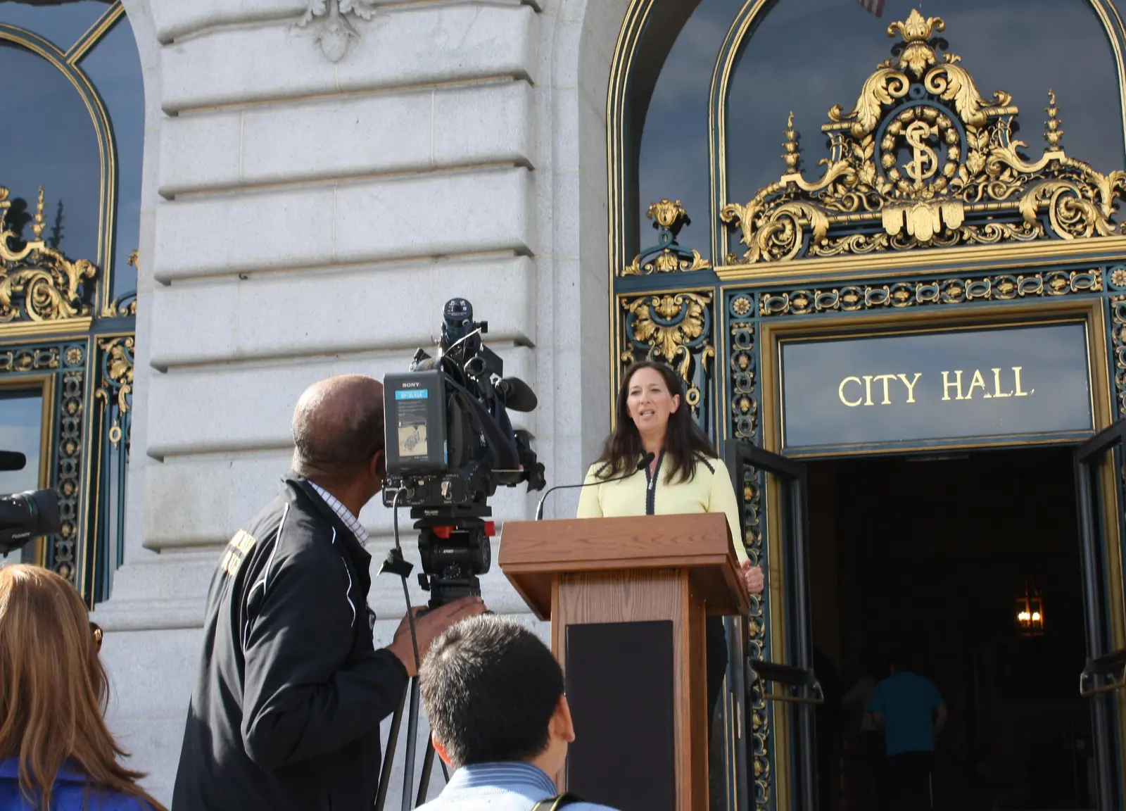 Nicole Ozer speaks at a podium outside City Hall, while several cameras capture her speech.