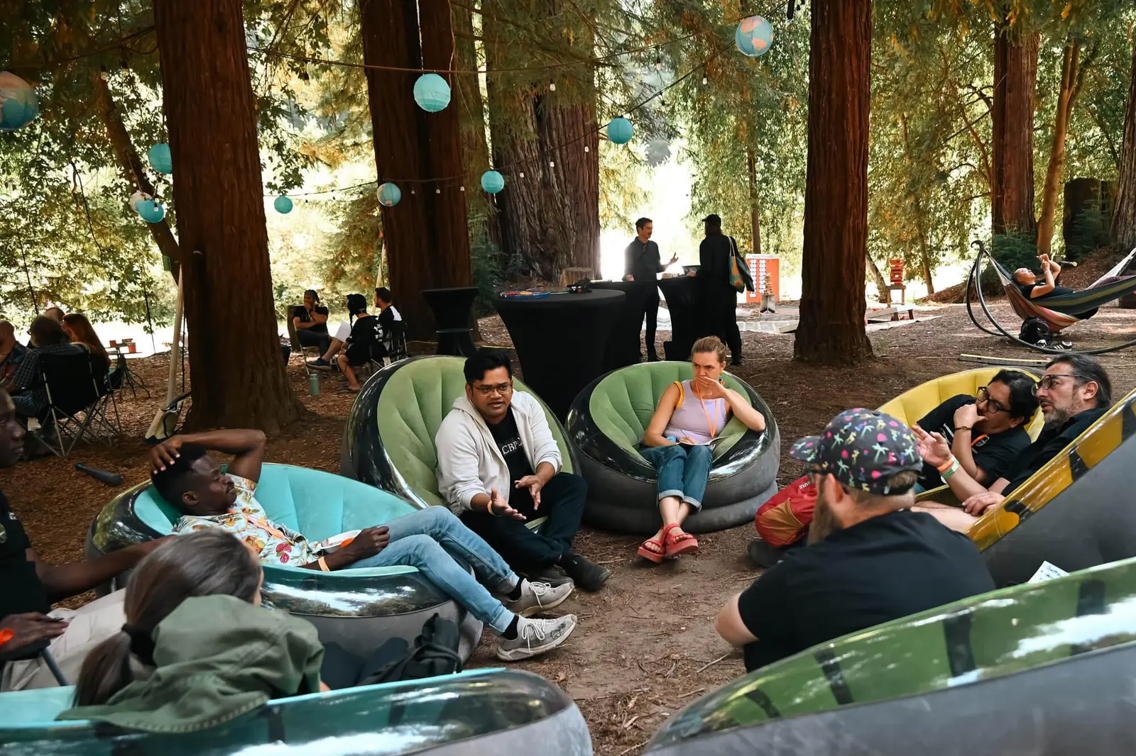 DWeb Camp attendees relaxing and conversing in inflatable lounge chairs among tall redwood trees, with teal paper lanterns strung overhead and a hammock visible in the background.