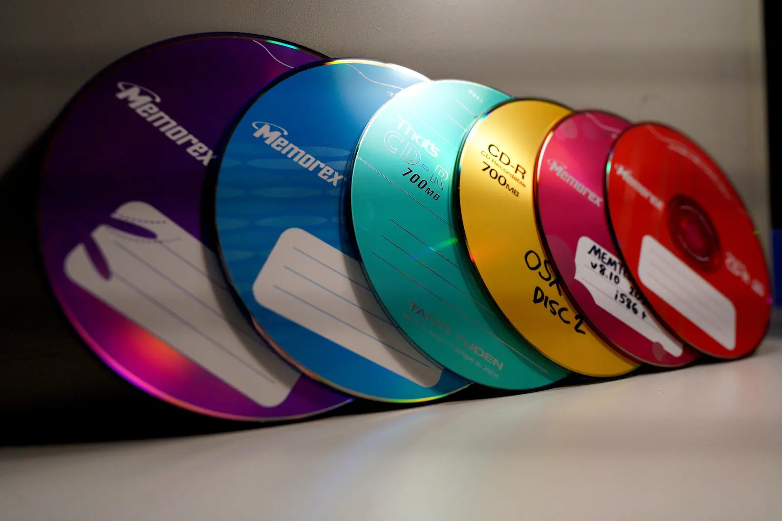 Six CD-Rs leaning against a beige backdrop. The photo is taken at an angle so that it kind of looks like I have a purple recordable laserdisc, which I sadly do not.

Purple, blue, pink (imitation orange), and red are Memorex, green is from Japanese brand "That's", and the yellow (actually gold) is Imation.

More banding across the photo.