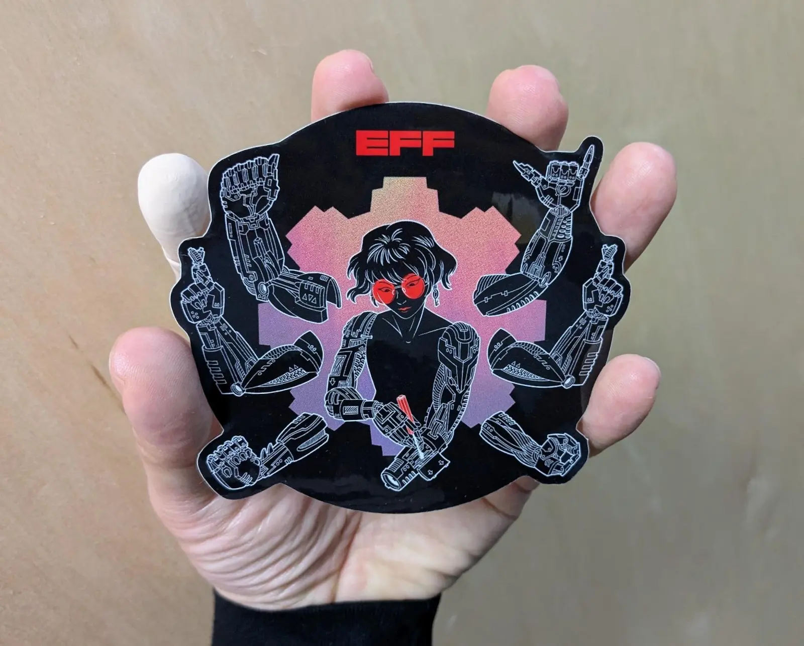 A hand holds a circular black sticker featuring a person with red glasses adjusting six mechanical arms with ASL signs.