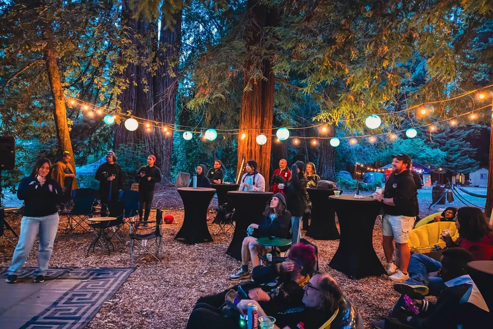 DWeb Camp attendees socializing at night among redwood trees, lit by warm string lights and glowing paper lanterns. People stand and sit around cocktail tables in a festive outdoor atmosphere.