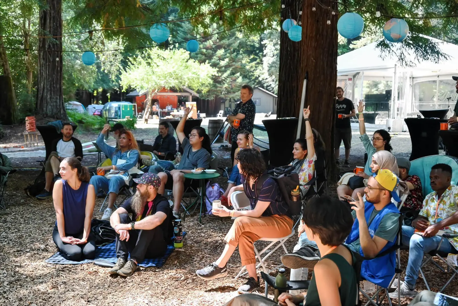 A diverse group of DWeb Camp attendees seated outdoors among redwood trees during a session, with several people raising their hands. Teal paper lanterns hang overhead, and a white event tent is visible in the background.