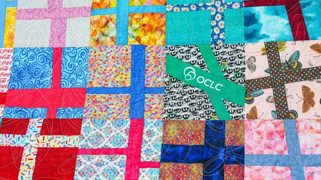 Close-up of a colorful patchwork quilt made of bright, patterned fabric squares and cross-shaped strips; a green strip includes the white OCLC logo.