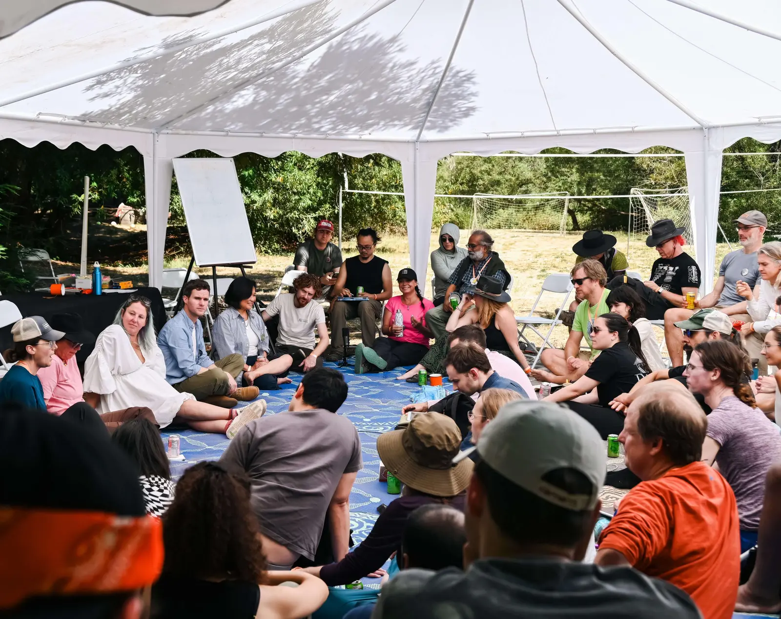 A large group of DWeb Camp attendees seated in a circle on a patterned rug inside a white open-sided tent, engaged in a structured group discussion, with a flipchart easel and sunny outdoor landscape visible behind them.