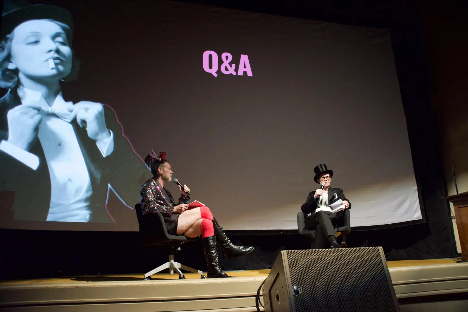 Chris Freeland and Denah Johnston sit onstage in the Internet Archive's great room during a Q&amp;A, in front of a large projection screen displaying "Q&amp;A" in pink text alongside a black-and-white film still. Chris wears a sparkly jacket, red stockings, and a small feathered hat; Denah wears a black top hat and blazer, both holding microphones.