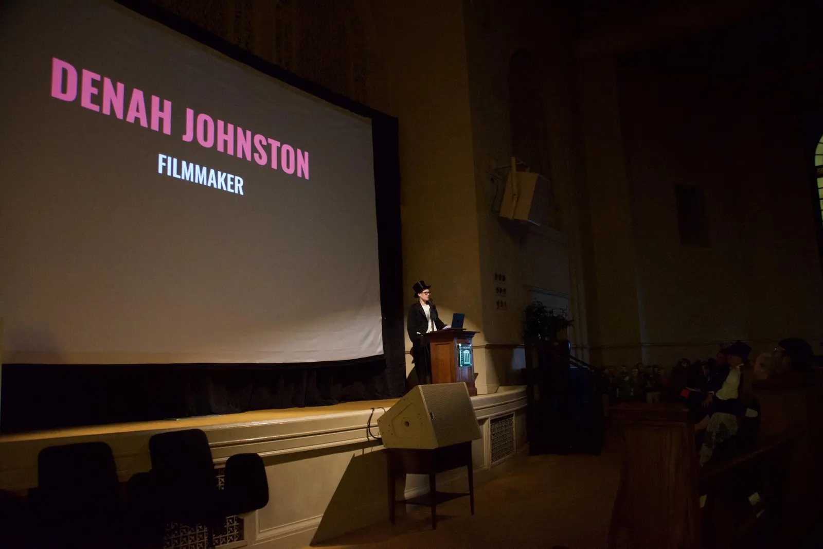 Filmmaker Denah Johnston stands at a lectern on the Internet Archive's great room stage. A large projection screen behind them reads "Denah Johnston, Filmmaker" in pink and white text.