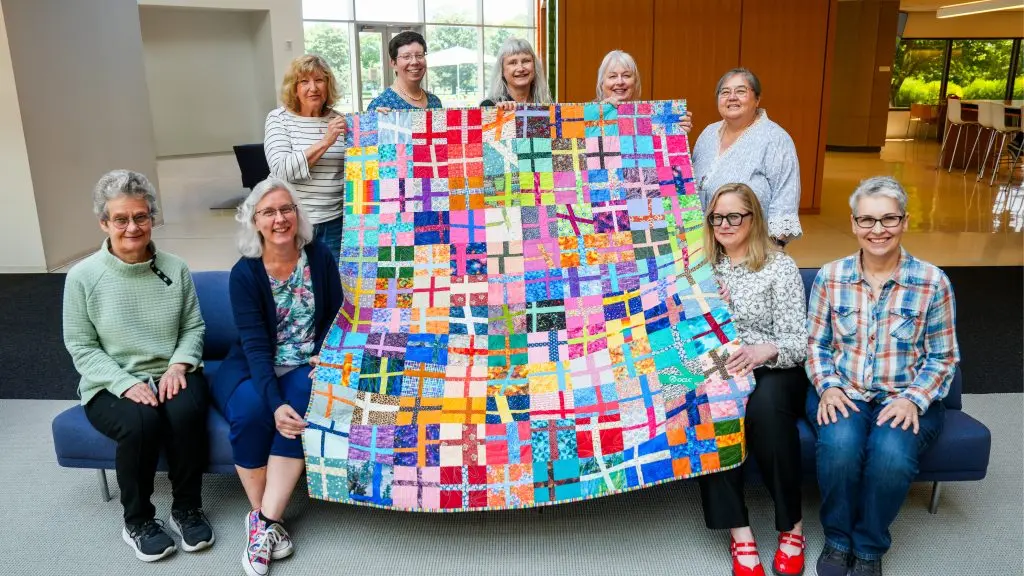 Nine people in a bright lobby pose with a large, colorful patchwork quilt featuring a grid of multicolored fabric squares; four stand behind holding the quilt Four people sit on a blue bench in front and five stand behind the bench